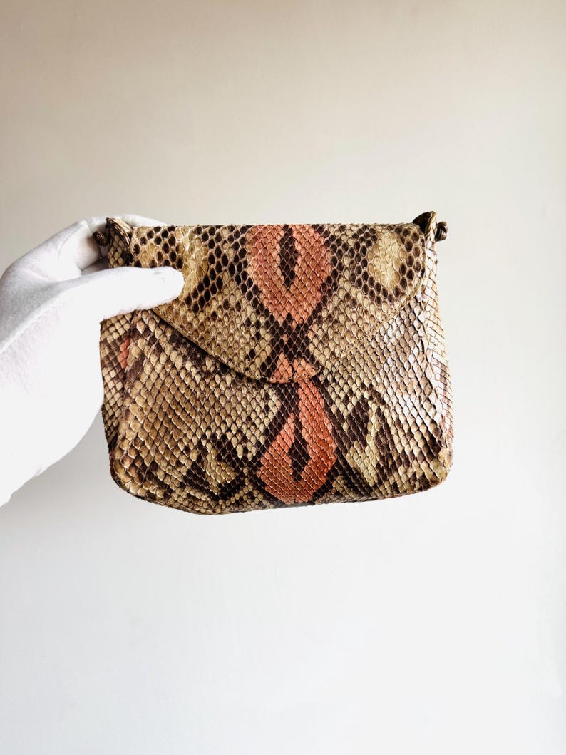 K&ouml;nnte beinhalten: Eine kleine, rechteckige Clutch mit einem Schlangenmuster in Braun-, Beige- und Pfirsicht&ouml;nen. Die Clutch hat einen Klappenverschluss und ein subtiles, strukturiertes Aussehen. Das Gesamtdesign ist kompakt und stilvoll, geeignet f&uuml;r das Tragen von Essentials.