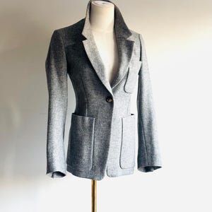 May include: A gray blazer with a tailored fit, displayed on a mannequin. The blazer features a notched lapel, two front pockets, and a single-button closure. The fabric appears to be a textured wool blend.