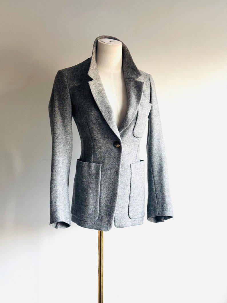 May include: A gray blazer with a tailored fit, displayed on a mannequin. The blazer features a notched lapel, two front pockets, and a single-button closure. The fabric appears to be a textured wool blend.