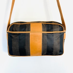 May include: A brown and black striped shoulder bag with a tan leather flap and a gold metal Fendi logo. The bag has a long tan leather strap.
