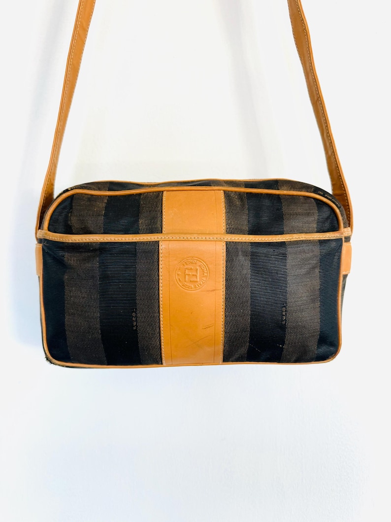 May include: A brown and black striped shoulder bag with a tan leather flap and a gold metal Fendi logo. The bag has a long tan leather strap.