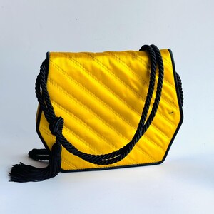 May include: A yellow satin handbag with a black rope handle and tassel. The bag has a quilted design with diagonal stitching. The edges are outlined in black. The bag is a unique hexagonal shape.