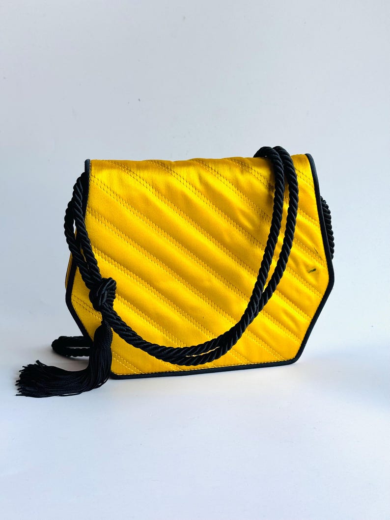 May include: A yellow satin handbag with a black rope handle and tassel. The bag has a quilted design with diagonal stitching. The edges are outlined in black. The bag is a unique hexagonal shape.