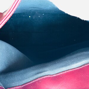 May include: Open view of a burgundy leather handbag. The interior is lined with a dark blue fabric. A silver-colored chain is visible on the right side. The bag's edges are neatly stitched.