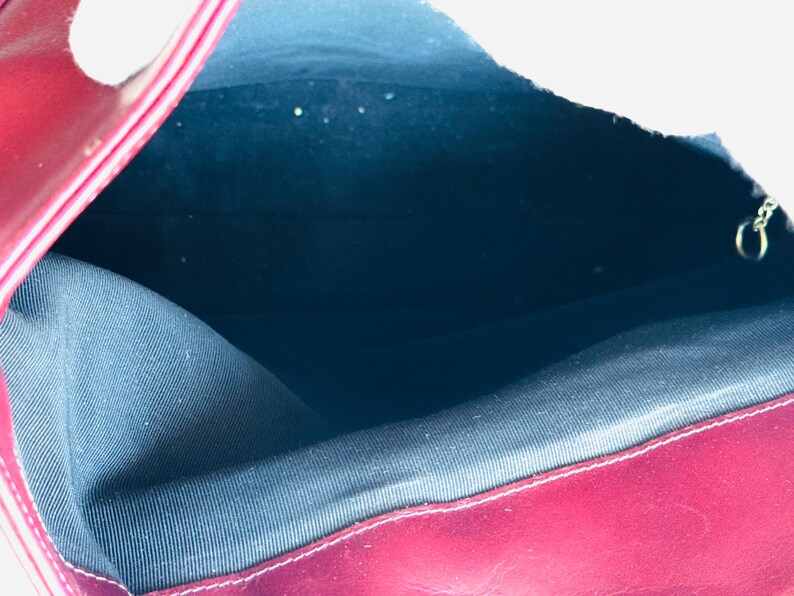 May include: Open view of a burgundy leather handbag. The interior is lined with a dark blue fabric. A silver-colored chain is visible on the right side. The bag's edges are neatly stitched.