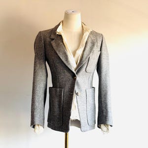 May include: A gray blazer with two front pockets and a single-button closure. The blazer is displayed on a mannequin, revealing a white collared shirt underneath. The jacket has long sleeves and a tailored fit, suggesting a classic style.