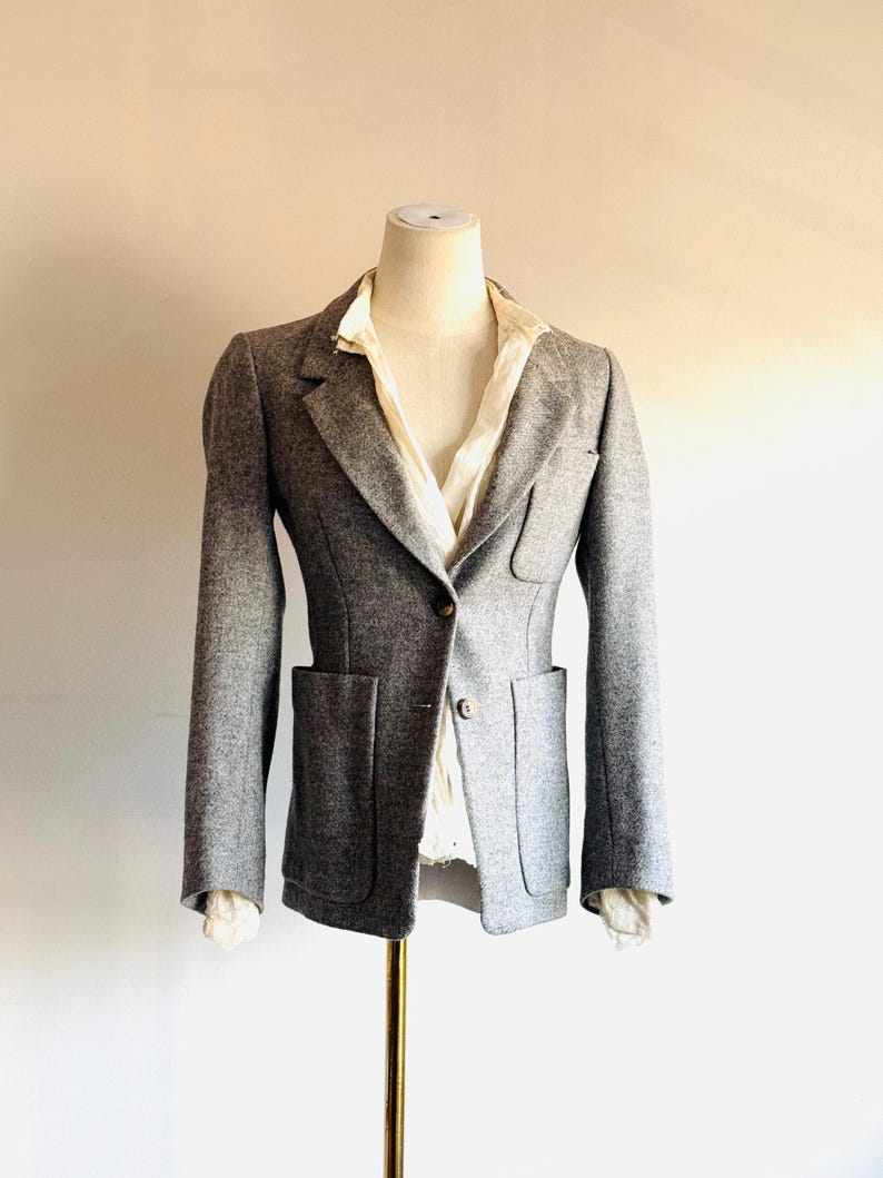 May include: A gray blazer with two front pockets and a single-button closure. The blazer is displayed on a mannequin, revealing a white collared shirt underneath. The jacket has long sleeves and a tailored fit, suggesting a classic style.