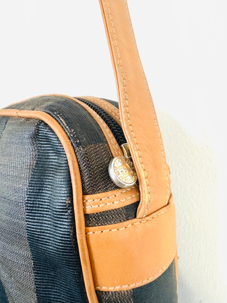 May include: A brown leather shoulder strap with a black and brown patterned bag. The bag has a zipper closure and a gold metal button with the letter "F" on it.
