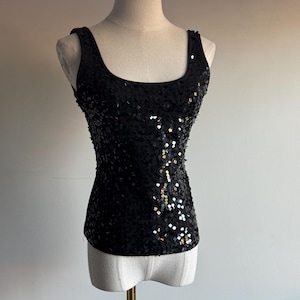 May include: A black sequined tank top with a scoop neckline. The top is displayed on a mannequin and features a fitted silhouette. The sequins create a shimmering effect, reflecting light. The garment is sleeveless and suitable for evening wear.