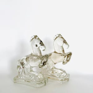 May include: A pair of clear glass horse bookends. The horses are standing on their hind legs with their front legs raised. The bookends are on a white background.