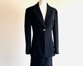 May include: A black two-piece suit displayed on a mannequin. The suit includes a blazer with a single gold button and a matching skirt. The fabric has a textured appearance. The suit is displayed on a gold stand against a white background.