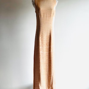 May include: A sleeveless, floor-length peach-colored dress with a textured pattern. The dress has a boat neckline and a straight silhouette. It is displayed on a mannequin.