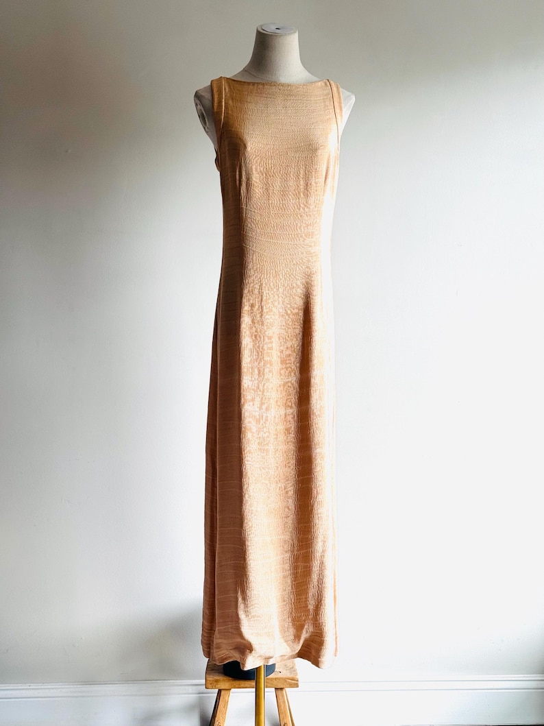 May include: A sleeveless, floor-length peach-colored dress with a textured pattern. The dress has a boat neckline and a straight silhouette. It is displayed on a mannequin.