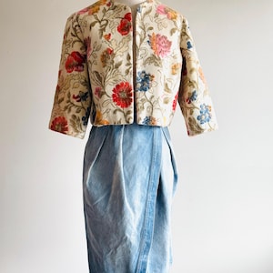 May include: A cream-colored, embroidered jacket with a floral pattern in red, pink, blue, and yellow. The jacket has three-quarter sleeves and is paired with a light blue denim skirt. The outfit is displayed on a mannequin.