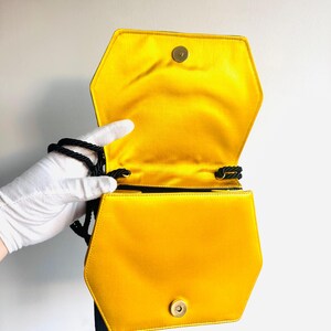 May include: Hexagonal yellow satin handbag with a black rope strap and tassel. The bag has a gold-colored clasp and a bright yellow interior. The bag is open, revealing the interior lining.