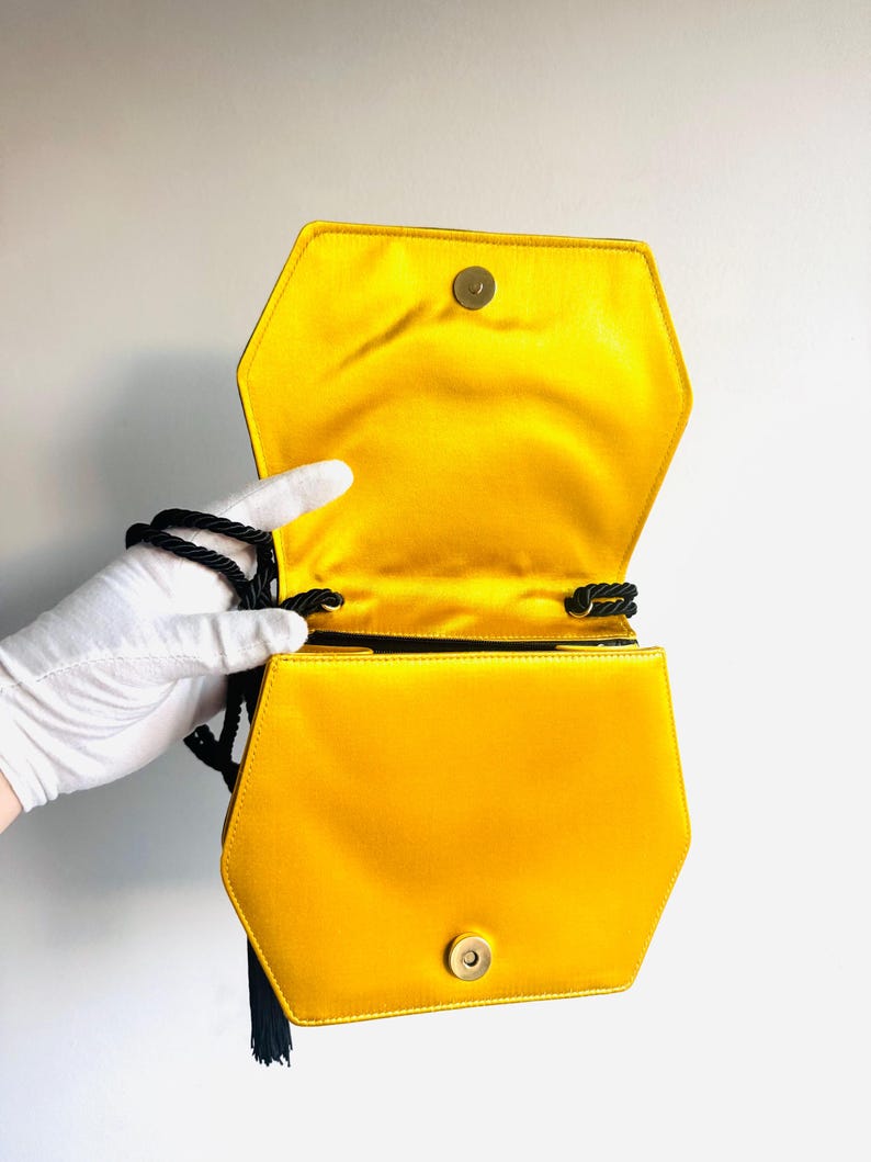 May include: Hexagonal yellow satin handbag with a black rope strap and tassel. The bag has a gold-colored clasp and a bright yellow interior. The bag is open, revealing the interior lining.