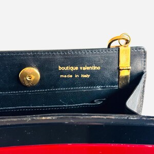 Może przedstawiać: Zbliżenie wnętrza czarnej sk&oacute;rzanej torebki ze złotymi elementami. Widoczny jest napis "boutique valentino made in Italy". Torba ma czerwono-biały pasek.