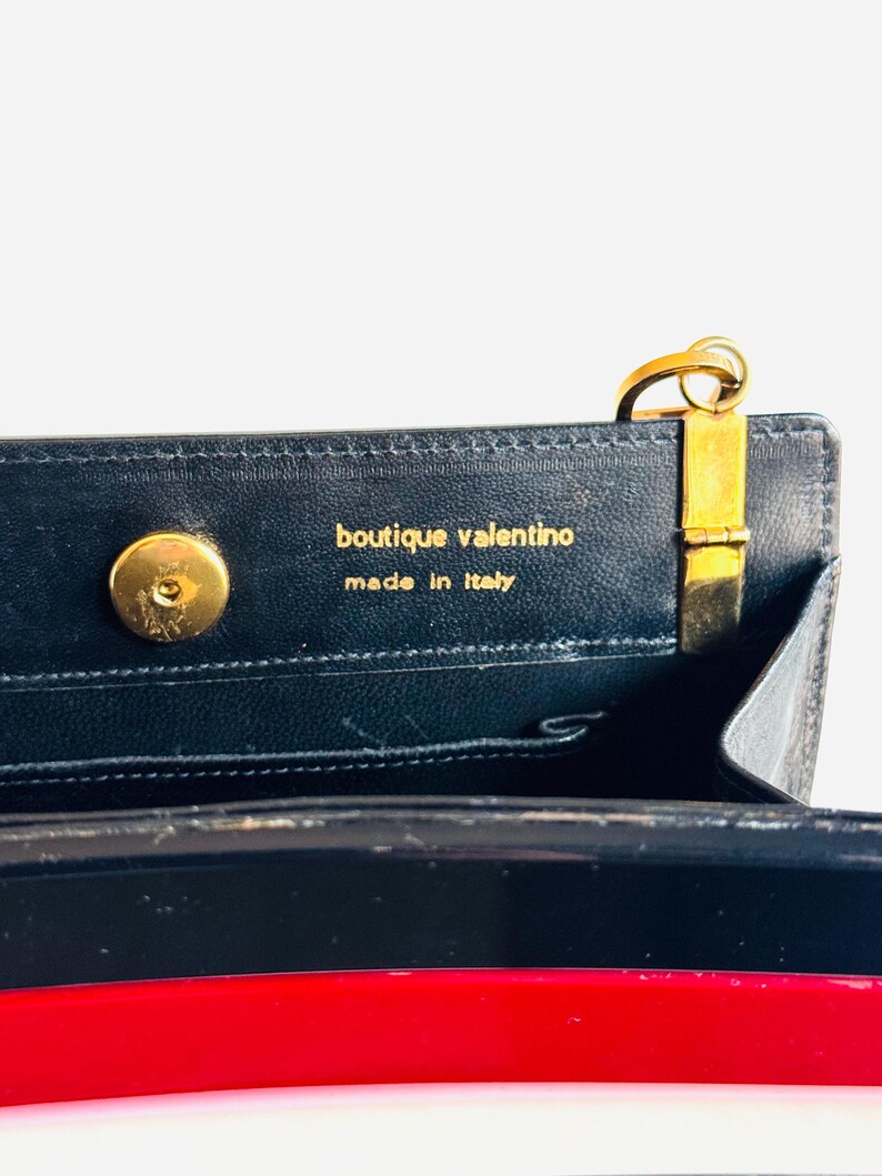 Może przedstawiać: Zbliżenie wnętrza czarnej sk&oacute;rzanej torebki ze złotymi elementami. Widoczny jest napis "boutique valentino made in Italy". Torba ma czerwono-biały pasek.