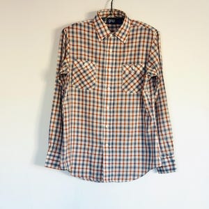 May include: A long-sleeved plaid shirt in shades of brown, blue, and white, with two chest pockets. The shirt is hanging on a decorative gold-colored hook. The brand name "gap" is visible on the label.