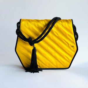 May include: A yellow quilted handbag with a black rope handle and trim. The bag has a geometric shape and a black tassel. The bag is made of a shiny fabric.