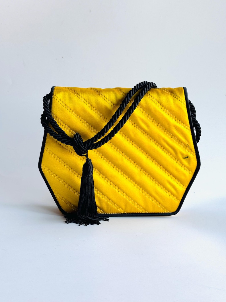 May include: A yellow quilted handbag with a black rope handle and trim. The bag has a geometric shape and a black tassel. The bag is made of a shiny fabric.