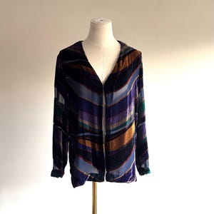 May include: A long-sleeved, open-front velvet jacket with a V-neck. The jacket features a striped pattern in shades of purple, blue, brown, and green. The sleeves have a slight ruffle at the cuffs. The jacket is displayed on a mannequin.