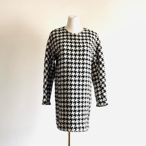 May include: A black and white houndstooth pattern dress with long sleeves. The dress is displayed on a mannequin. The dress has a round neckline and a slightly flared shape. The fabric appears to be a textured knit.