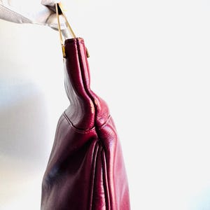 May include: A burgundy leather handbag with gold-toned handles. The bag is held up by a gloved hand, showcasing its shape and color. The bag's design features a curved silhouette and a subtle sheen.