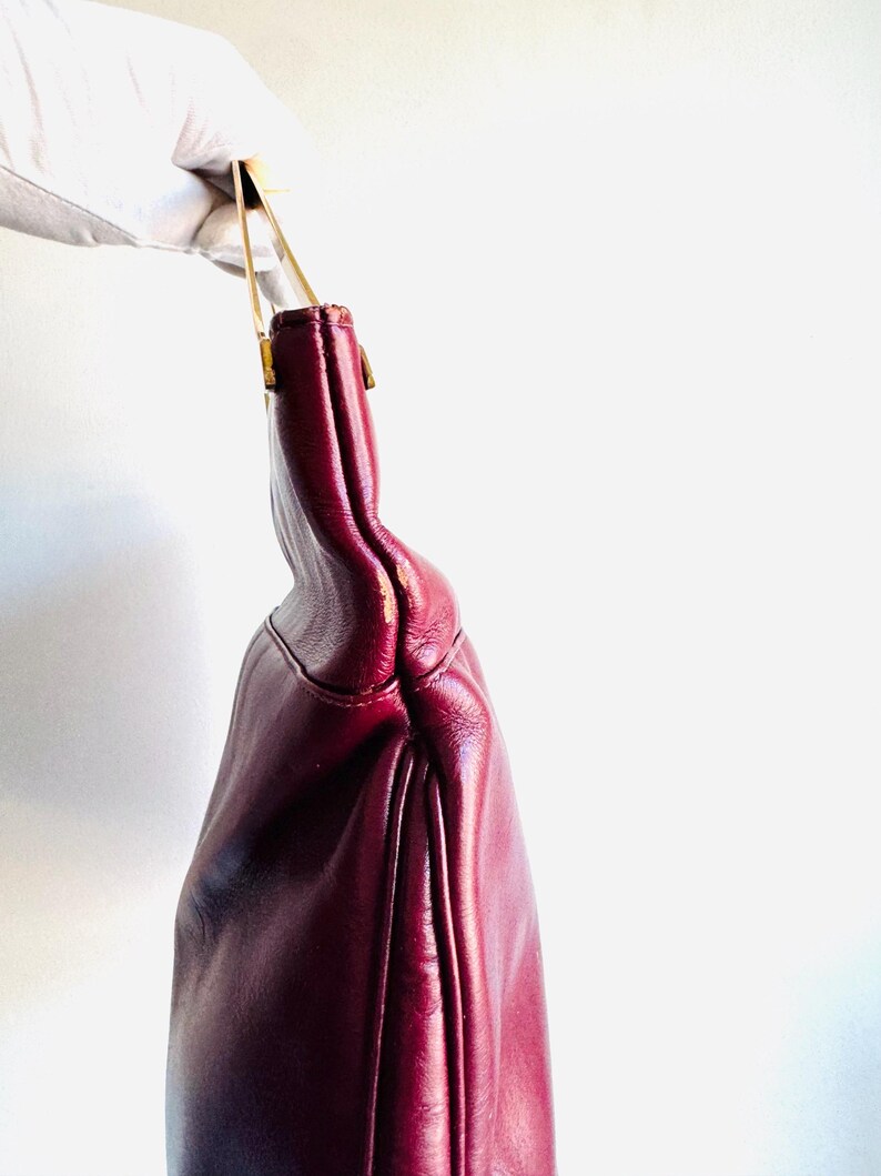 May include: A burgundy leather handbag with gold-toned handles. The bag is held up by a gloved hand, showcasing its shape and color. The bag's design features a curved silhouette and a subtle sheen.