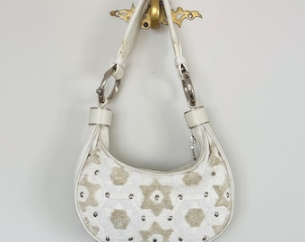 Puede incluir: Bolso de hombro blanco con forma de media luna. El bolso presenta un patrón decorativo de adornos beige y plateados. Tiene una correa de cuero blanco y herrajes metálicos plateados. El bolso está colgado de un gancho dorado.