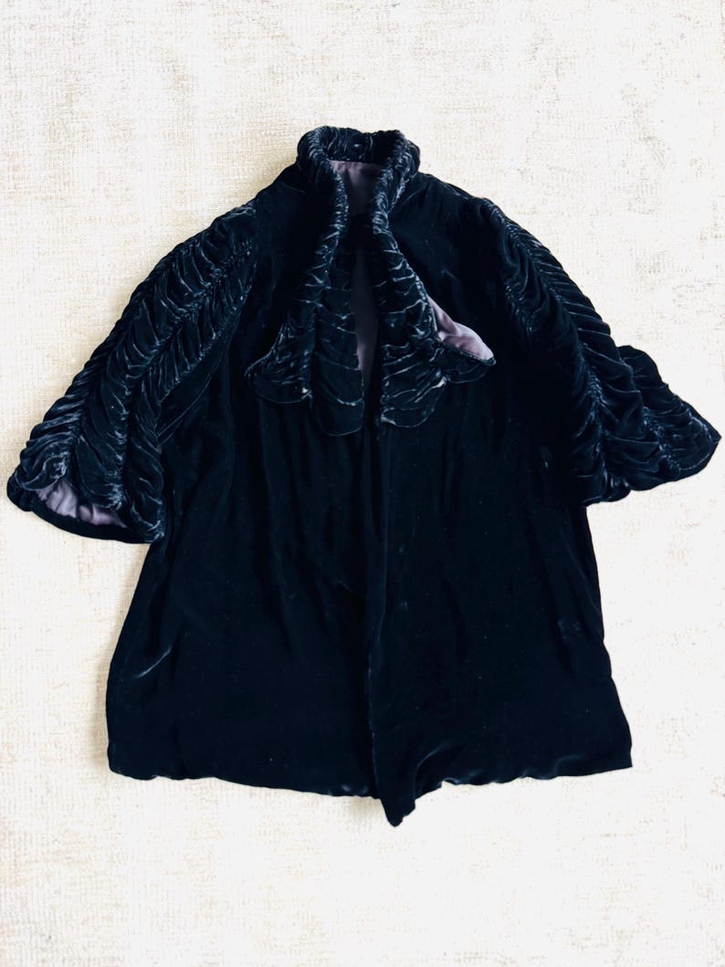 K&ouml;nnte beinhalten: Eine schwarze Samtjacke mit einem R&uuml;schenkragen und R&uuml;schen&auml;rmeln. Die Jacke ist mit einem violetten Stoff gef&uuml;ttert.