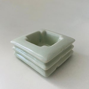 May include: A light green ceramic ashtray with a square shape and three layers. The ashtray is designed with a raised rim around the top layer.