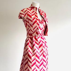 May include: A vintage red and cream-colored dress and matching jacket set. The dress features a chevron pattern with a high neckline and a fitted waist. The jacket has short sleeves and a matching chevron pattern. The outfit is displayed on a mannequin.