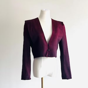 May include: A burgundy cropped blazer with a V-neckline and long sleeves. The blazer has three small, dark buttons down the front and is displayed on a white mannequin. The fabric has a subtle sheen.