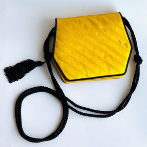 May include: A yellow, hexagonal handbag with black trim and a black rope strap. The bag has a diagonal stitched pattern and a black tassel. The bag is on a white background.