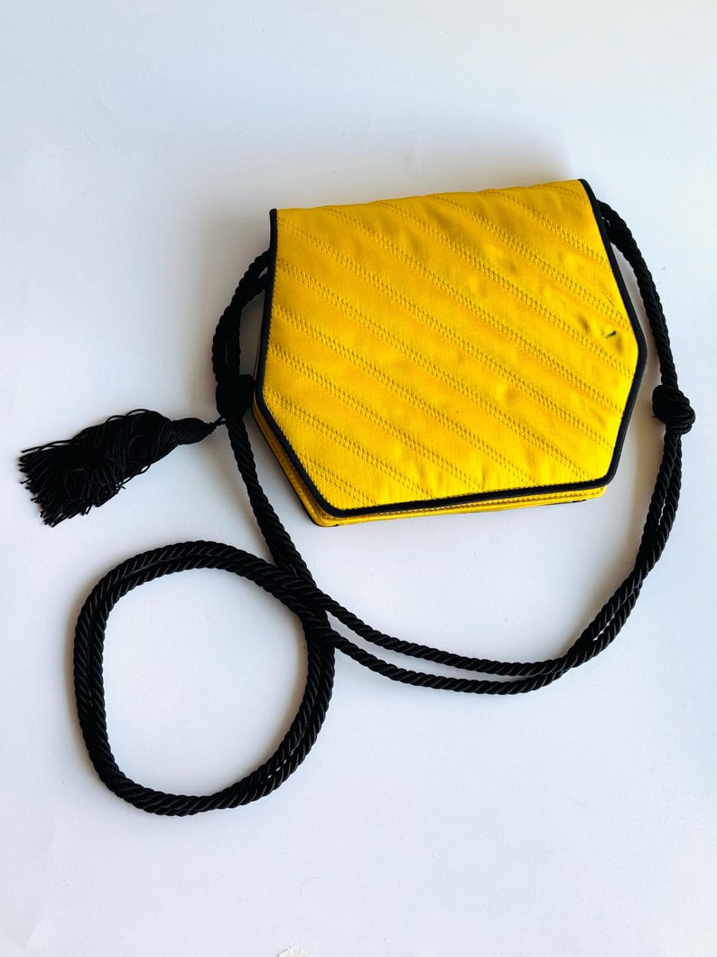 May include: A yellow, hexagonal handbag with black trim and a black rope strap. The bag has a diagonal stitched pattern and a black tassel. The bag is on a white background.