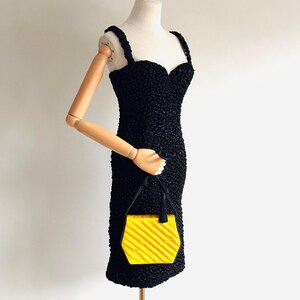 May include: A black textured dress with a sweetheart neckline and spaghetti straps is displayed on a mannequin. A small, yellow, geometric handbag with a black handle is held by the mannequin's arm. The handbag has a ridged design.