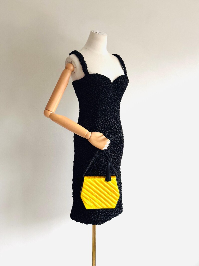 May include: A black textured dress with a sweetheart neckline and spaghetti straps is displayed on a mannequin. A small, yellow, geometric handbag with a black handle is held by the mannequin's arm. The handbag has a ridged design.