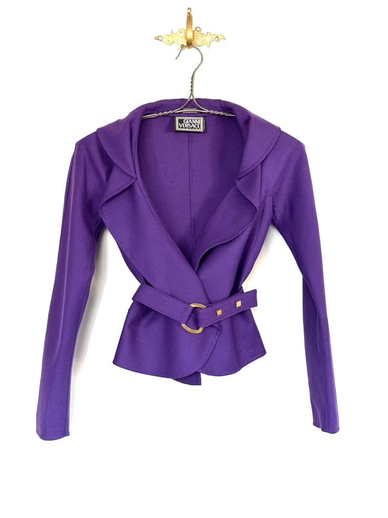 Pode incluir: Um casaco roxo vibrante com um design envolvente e um cinto a condizer com fivela dourada. O casaco tem mangas compridas e gola entalhada. A etiqueta diz "Gianni Versace".