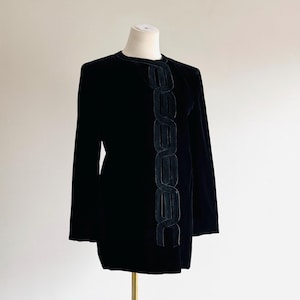 May include: Black velvet jacket with long sleeves and a decorative vertical design down the front. The design is made of a darker, possibly leather-like material. The jacket is displayed on a mannequin.