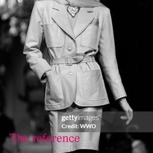 May include: A black and white image of a fashion model wearing a light-colored suit with a matching skirt and a wide-brimmed hat. The suit has a belted waist and large pockets. The model is wearing gloves and stockings.