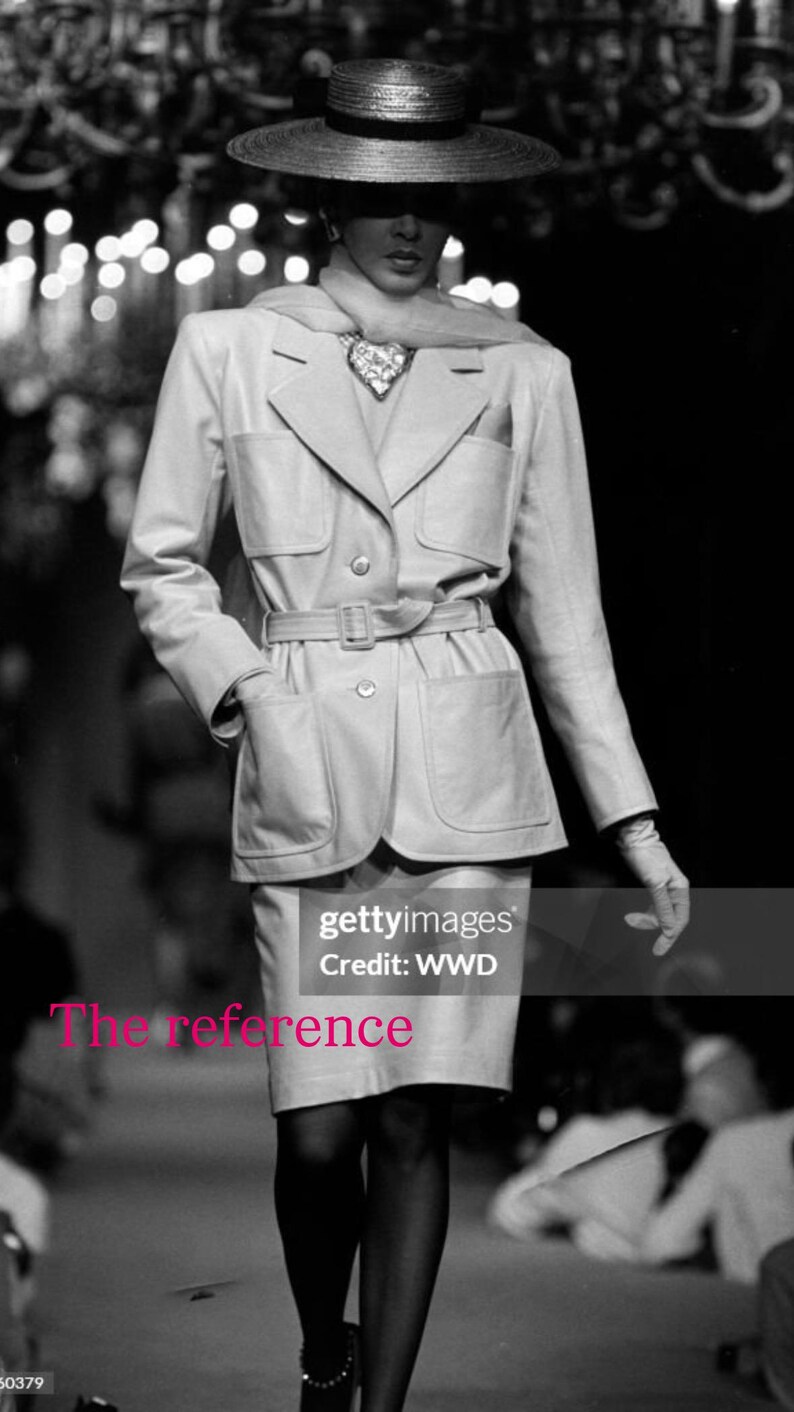 May include: A black and white image of a fashion model wearing a light-colored suit with a matching skirt and a wide-brimmed hat. The suit has a belted waist and large pockets. The model is wearing gloves and stockings.