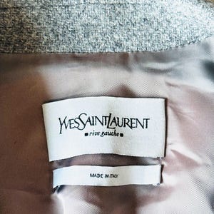 May include: Close-up of the interior of a gray blazer. The lining is a light brown color. Two white tags are visible, one with the text "Yves Saint Laurent rive gauche" and the other with "MADE IN ITALY".