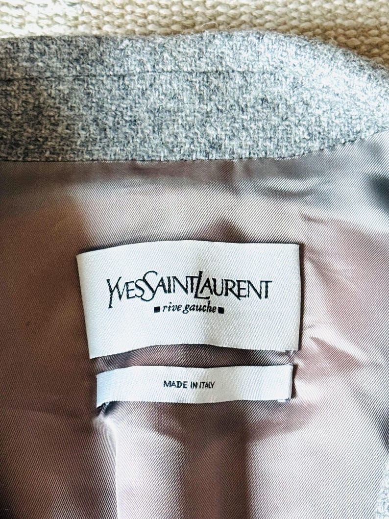 May include: Close-up of the interior of a gray blazer. The lining is a light brown color. Two white tags are visible, one with the text "Yves Saint Laurent rive gauche" and the other with "MADE IN ITALY".