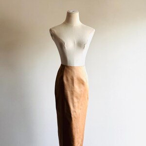 May include: A tan pencil skirt displayed on a mannequin. The skirt is a solid color with a slight sheen, and it reaches to the knees. The mannequin is a neutral color, and the background is a plain white wall.