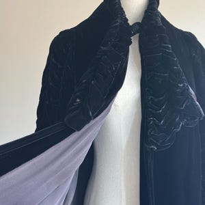 K&ouml;nnte beinhalten: Ein schwarzer Samtmantel mit einem R&uuml;schenkragen und einer langen, flie&szlig;enden Silhouette. Der Mantel ist mit einem dunkelgrauen Stoff gef&uuml;ttert.