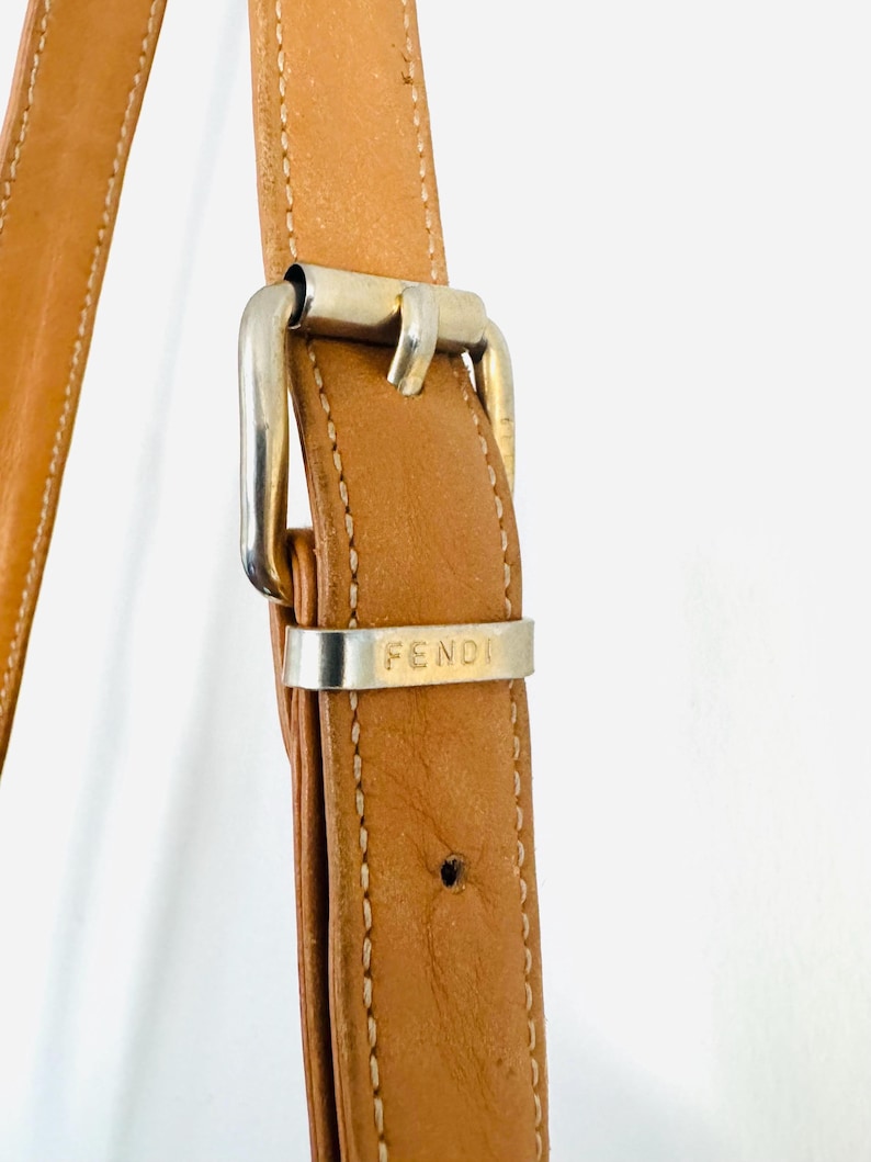 May include: Close-up of a brown leather shoulder strap with a silver buckle. The buckle has the word "FENDI" engraved on it.