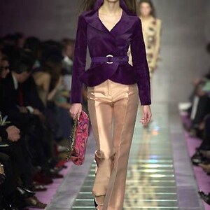 Pode incluir: Modelo numa passarela vestindo um casaco de veludo roxo com cinto a condizer e cal&ccedil;as castanhas claras. O casaco tem uma gola larga e uma peplum. As cal&ccedil;as s&atilde;o largas. A modelo carrega uma bolsa com padr&atilde;o rosa e roxo.