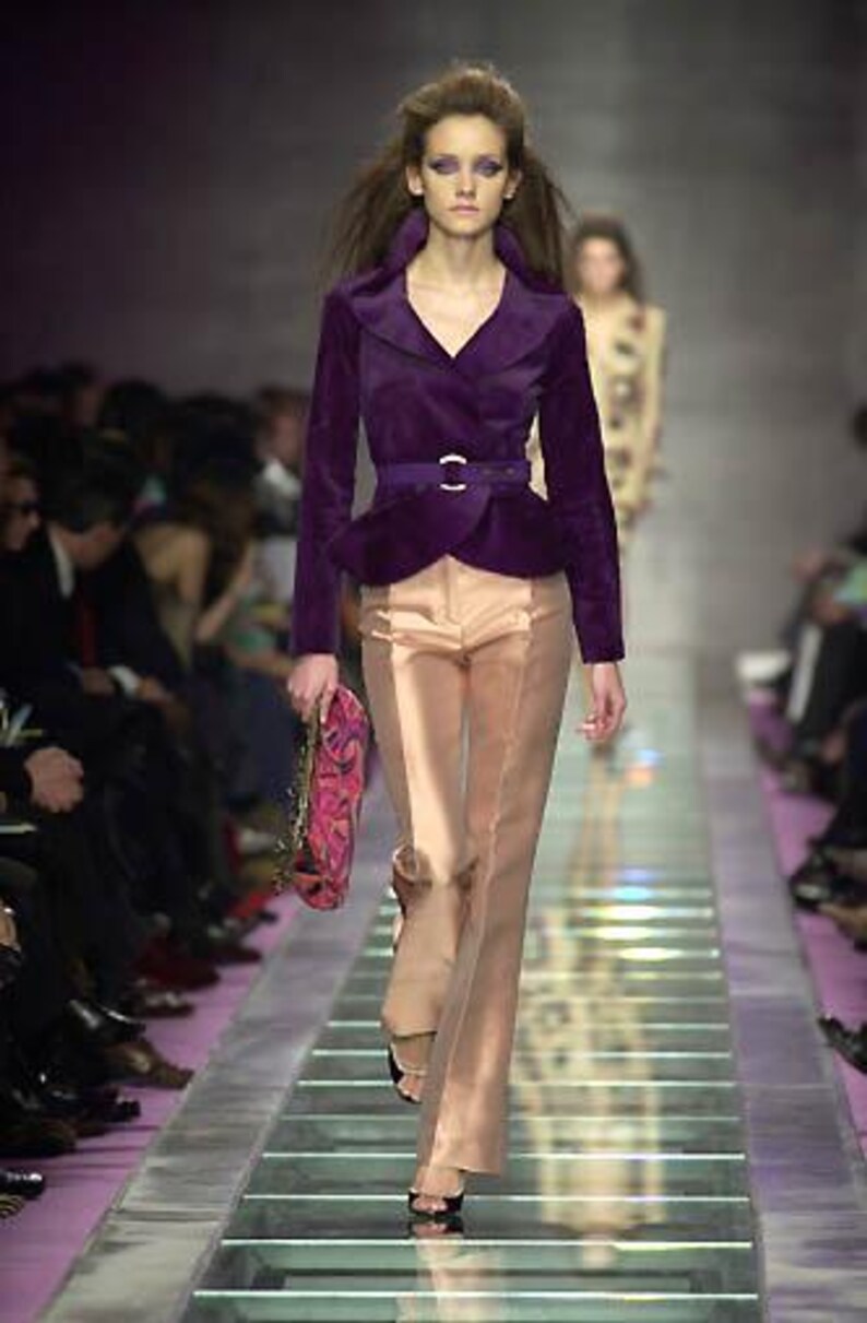 Pode incluir: Modelo numa passarela vestindo um casaco de veludo roxo com cinto a condizer e cal&ccedil;as castanhas claras. O casaco tem uma gola larga e uma peplum. As cal&ccedil;as s&atilde;o largas. A modelo carrega uma bolsa com padr&atilde;o rosa e roxo.