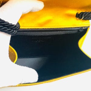 May include: Open view of a yellow satin clutch with a black rope handle. The interior is black with a small zipper pocket. The words "MADE IN ITALY" are visible.